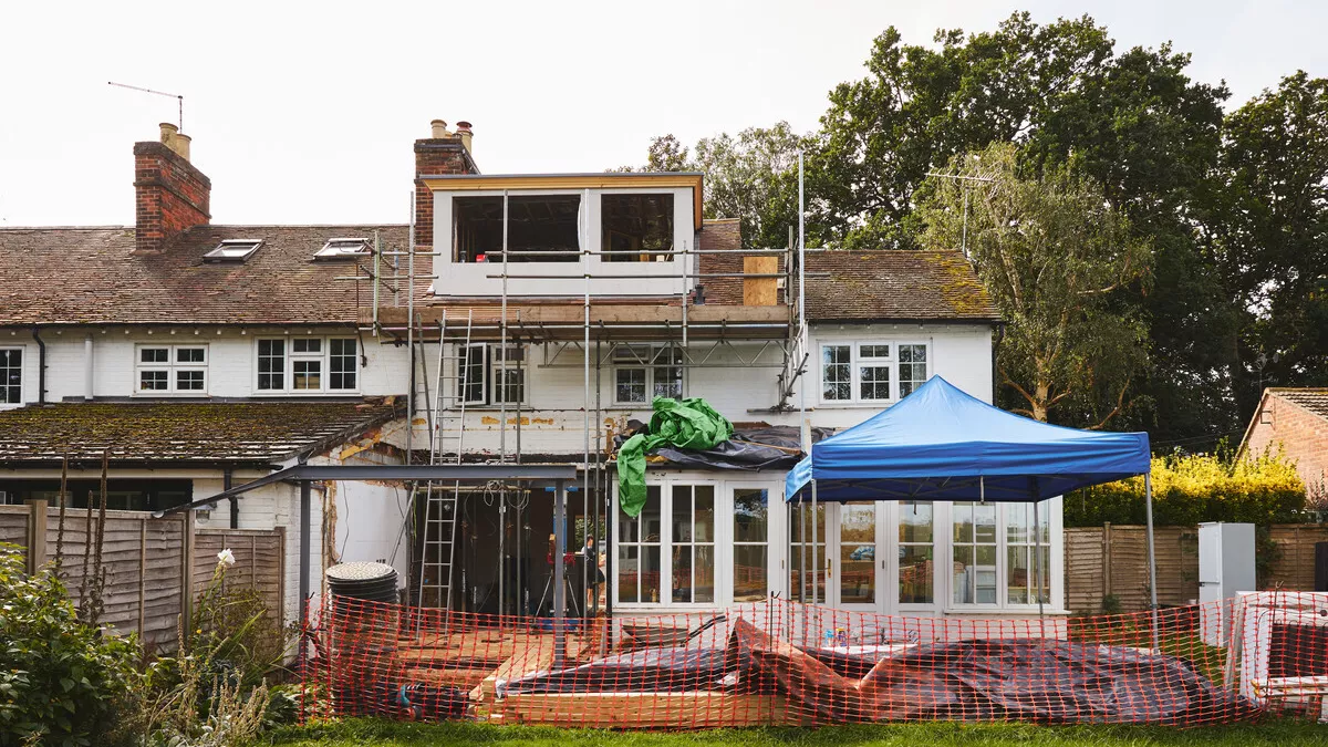 House under construction with scaffolding, blue tent, and landscaping in progress.