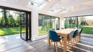 Bright dining area with a wooden table, blue chairs, and large glass windows overlooking a garden.