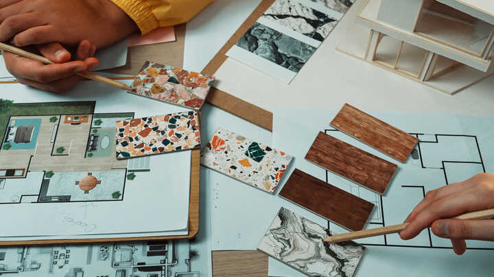 Hands holding brushes over design plans and material samples on a table.
