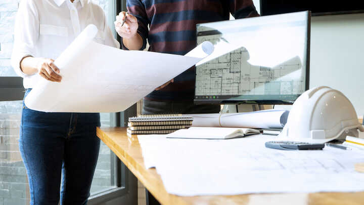 Two people reviewing blueprints on a table with a computer and construction helmet nearby.