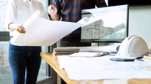 Two people reviewing blueprints on a table with a computer and construction helmet nearby.