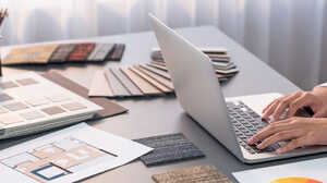 A person types on a laptop at a desk with papers and colored pencils.