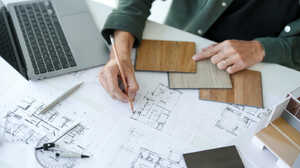 Designer marking up a floor plan with timber and flooring samples laid out alongside a laptop and scale rule