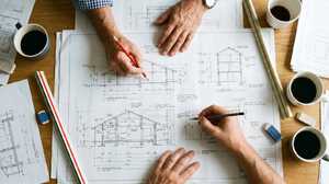 Two designers comparing elevation and section drawings across a desk of coffee cups and drawing instruments