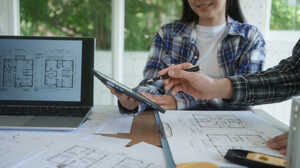 wo people discussing floor plans over a tablet, with a laptop and printed architectural drawings on the desk.