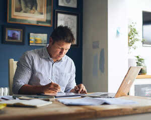 A man working at a desk, writing on papers with a laptop in front of him.
