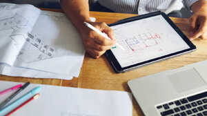 A person uses a stylus on a tablet while studying printed papers and a laptop.
