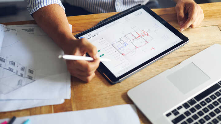 Person using a tablet and laptop, reviewing architectural drawings on a wooden desk.