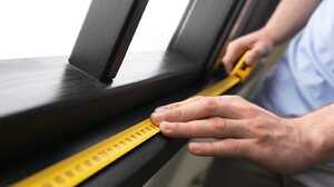 Hands measuring a dark timber window frame with a yellow tape measure